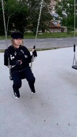 A young person wearing a black baseball cap, a black hoodie with white star details, and black pants is sitting on a swing set. They are also wearing black gloves and white-toed sneakers. The person is in motion, swinging back and forth. The swing set is located in an outdoor area with a paved ground. In the background, there are trees, a sidewalk, and a building. The sky is overcast.