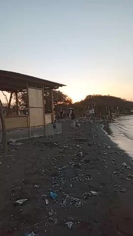 A group of people are walking along a dirty beach near some small structures. The sky is clear and the sun appears to be setting, casting a warm glow over the scene. The beach is covered in trash, and there are several small buildings constructed of wood and corrugated metal. The water is calm and appears to be gently lapping at the shore.