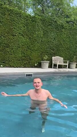 A shirtless man stands waist-deep in a swimming pool. He has a buzz cut and wears a gold necklace and striped swim trunks. He looks directly at the camera. In the background, a tall, manicured hedge lines the edge of the pool area. To the right, there is a wooden bench and two potted plants. The water in the pool is clear and slightly blue.