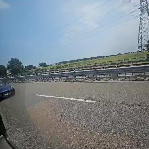 The video begins with a blue car in the left lane of a highway. The perspective then shifts to the interior of another vehicle, showing the blue car in the rearview mirror as it passes under a bridge. The camera then returns to an exterior view, showing the blue car ahead in the left lane of the highway. The blue car continues to move forward, with the landscape of trees and fields visible on either side of the road. The video ends with the blue car still visible in the distance on the highway.
