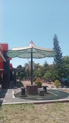 The video pans across an outdoor area with a clear blue sky. In the foreground, there is a patch of dry grass and a paved walkway with a floral design. A large umbrella-shaped canopy with a white fabric cover and a brown pole provides shade over a circular seating area with three dark-colored benches arranged around a central dark cylindrical base. Beyond this seating area, there is more greenery, including trees and bushes. Further back, a tall, slender pine tree stands out. In the background, a building with a red section is visible to the left. Several cars are parked under a covered area. A lamppost with a white spherical light fixture stands in the middle ground. The overall environment appears to be a well-maintained public space or park, possibly on a sunny day.