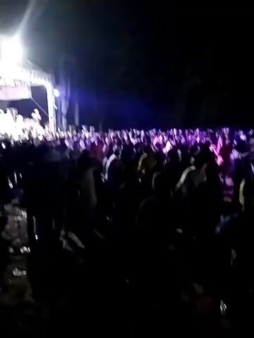 The media file depicts a nighttime outdoor concert in Krebet, Indonesia. A large crowd of people are gathered in front of a brightly lit stage. The stage is adorned with a metal truss structure and appears to be set up for a musical performance, with instruments and equipment visible. Several individuals are on stage, presumably the performers, with one person speaking into a microphone. The audience is silhouetted against the stage lights, with many individuals raising their hands in the air, suggesting an energetic and engaged atmosphere. The ground in front of the stage is uneven and appears to be dirt or gravel, with some debris scattered about. The overall mood of the scene is one of excitement and festivity, typical of a live music event. There is some visible text on banners on the stage, including "KOMORENSA". The time of day is clearly night due to the darkness of the surroundings and the prominence of the stage lighting. No weather information is available.