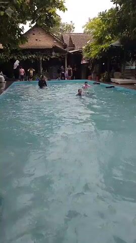 The video shows a swimming pool with several people in it. In the background, there is a building with a thatched roof and trees. The people in the pool are swimming and playing. The water is clear and blue. The sun is shining and the weather appears to be warm.