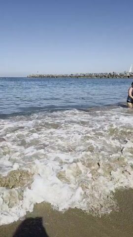 The video shows a close-up, low-angle shot of gentle waves washing onto a sandy beach. In the foreground, the water is clear and shallow, revealing the sandy bottom with small rocks. White foam from the waves creates a frothy texture as it recedes. A dark shadow, likely from the camera person, is visible on the lower left of the frame. In the background, the ocean stretches out to the horizon under a clear blue sky. A long, rocky breakwater extends into the water from the right side of the frame, with a small structure or beacon at its far end. The overall scene is peaceful and serene, capturing the natural beauty of a coastline on a sunny day.