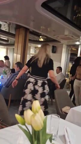 The video shows the interior of a restaurant where several women are dancing. The camera is positioned near a table set with a white tablecloth, glassware, and a vase of yellow tulips. The women are dancing in the open space near the tables; one is wearing a black top and a black-and-white floral skirt. Other people are seated at nearby tables. The restaurant has a modern design, with light-colored walls, dark chairs, and reflective surfaces. A man in a light-colored shirt sits at a table in the background.