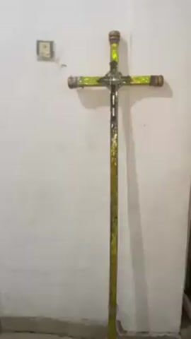 The video shows a standing cross with an ornate centerpiece. The cross is covered in a reflective, iridescent material that gives off green and gold hues. The centerpiece is a detailed, silver-colored design with intertwined letters. The cross itself appears to be constructed from a pipe-like material, with copper caps on the ends of the crossbars. The cross stands against a plain, off-white wall, near an electrical outlet.