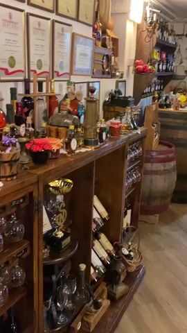 This video captures an interior view of a charming, rustic room, likely a wine shop or tasting area in Meterizi, Montenegro. The space is filled with wooden furnishings, including shelves, counters, and two prominent wooden barrels used as display tables.

Numerous bottles, possibly containing wine, spirits, or local produce, are neatly arranged on shelves and the barrel tops. Many wine glasses are visible, both on racks and resting on surfaces. The walls are decorated with several framed diplomas and awards, one clearly showing the word "DIPLOMA." Various decorative items, such as pomegranates, small floral arrangements, statues, and traditional scales, enhance the cozy atmosphere. A wooden wine rack holding several bottles is positioned in the foreground. The room features warm indoor lighting and a light-colored tiled floor. No people are present, and no specific activity is taking place, presenting a static view of the well-decorated space.