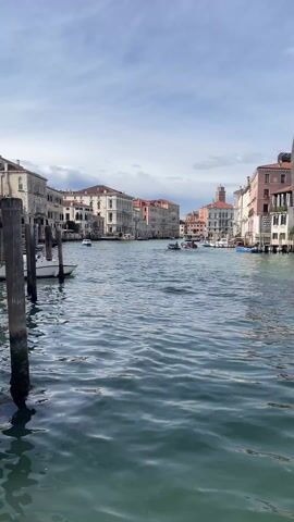 The video shows a view of the Grand Canal in Venice, Italy. The water is a dark blue-grey and has some ripples. On either side of the canal, there are tall buildings in a variety of architectural styles and colors, including white, cream, and red. There are boats moving on the canal and docked on the sides. The sky is overcast with grey and white clouds.