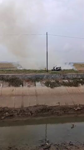 A motorcycle is parked on a dirt path next to a field. Smoke is rising from the field, obscuring the view of the horizon. The field appears to be dry, and there are patches of vegetation visible. A concrete canal runs along the bottom of the frame, filled with murky water. The sky is overcast.