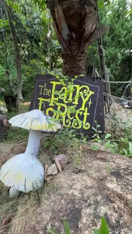 The video shows an outdoor scene, likely the entrance to a themed area called "The Fairy Forest," in Nam Nha Trang, Vietnam. In the foreground, a large, white artificial mushroom with yellow-painted accents on its cap is prominent, resting on the brown, earthy ground. Further back and slightly to the right, a dark sign with the words "The Fairy Forest" written in a bright, stylized yellow font is visible. Green vines are intertwined with the letters on the sign. Another, smaller artificial mushroom and tree stumps are visible in the background, amidst lush green foliage and trees. The camera slowly moves forward, capturing the whimsical details of this entrance. The natural lighting suggests it is daytime.