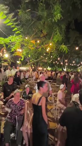 The video shows a crowded outdoor gathering at night. String lights are strung through trees, illuminating the scene. People are standing and sitting in rows of chairs, and some are walking around. The overall atmosphere appears festive, possibly a wedding or a similar celebration.