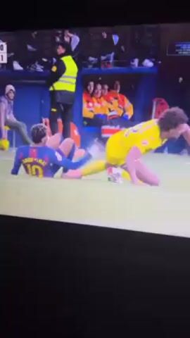 A video shows a soccer player wearing a blue and red uniform with the number 10 lying on the grass seemingly in pain. Another player wearing a yellow uniform is kneeling nearby. In the background, two people in orange and yellow jackets sit on a bench. An individual in a neon yellow vest stands nearby. The score on the screen reads "0-1".