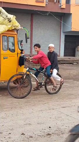 Two young boys are on a bicycle, with one boy pedaling and the other sitting on the back. They are in front of a yellow truck with colorful decorations. The boys appear to be smiling and enjoying themselves. The scene takes place outdoors on a street.