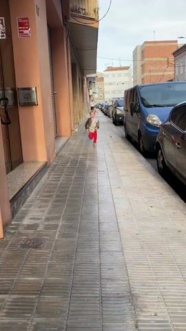 A young boy with blond hair runs down a narrow, tiled sidewalk bordered by buildings and parked cars. He is wearing a jacket, a dinosaur-patterned shirt, and red pants. He smiles widely as he runs toward the camera. As he reaches the camera, he jumps into the arms of a woman wearing a pink shirt and sunglasses perched on her head. She embraces him tightly.