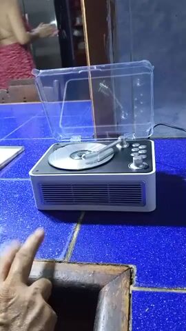 A person's hands are visible in the foreground of the video, interacting with a vintage-style record player. The record player is white with black accents and has a clear plastic lid that is open. The turntable is visible with a record on it. The background shows a person in a red and white outfit standing in front of a mirror and a kitchen area with stainless steel appliances. The record player is placed on a blue tiled surface. The person's hands are shown closing the lid of the record player.