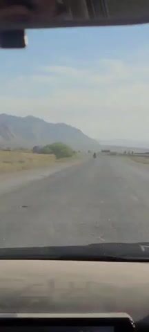 The view from inside a vehicle, looking out through the windshield, reveals a long, straight road stretching into the distance. The road is gray and appears somewhat worn. In the distance, a motorcycle is moving along the road, heading in the same direction as the camera. A vehicle is also farther down the road. To the right of the road, a group of people are standing beside what appears to be a barrier or edge of some kind. The landscape surrounding the road is arid, with mountains visible in the background under a sky that is predominantly blue with some wispy clouds. A small patch of green vegetation is visible to the left of the road.