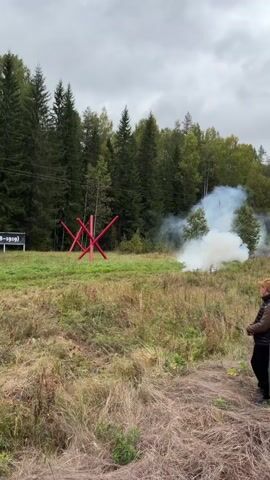 The video shows a forested area with a cloudy sky. In the foreground, there is dry grass and a person standing on the right side of the frame, looking towards the left. In the middle ground, there is a red, spiky obstacle resembling a caltrop. Behind the obstacle, a plume of white smoke is visible, suggesting a smoke bomb or pyrotechnic device has been detonated. To the left of the obstacle, there are several signs with Russian text. Further back, the trees are dense, creating a forest backdrop. The overall scene appears to be an outdoor event or demonstration, possibly related to a historical reenactment or a military-themed exhibit.