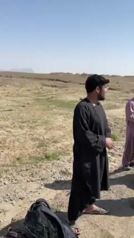The video shows a group of men standing beside a road in a desert landscape. In the first shot, two men are visible: one wearing a black robe and another wearing a light purple robe.  They are standing on a paved area near a dirt road, with a backpack at their feet. In the background, there are scrubby plants and a low hill.

The scene shifts to show more men standing near a light-colored car parked on the side of a paved highway. Some of the men are wearing traditional clothing, including head coverings.  The highway stretches into the distance, and mountains are visible on the horizon under a clear blue sky.  A person is seen kissing another.