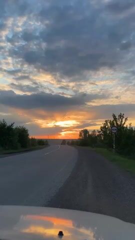 A car drives along a dark asphalt road at sunset. The sky is a mix of dark blue and grey clouds, with patches of golden light peeking through. The sun is visible just above the horizon, casting a warm glow on the surrounding landscape. The road is flanked by green bushes and trees on either side. A traffic sign indicating “No entry for motor vehicles” stands on the right side of the road. The reflection of the sunset can be seen on the hood of the car.