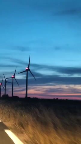 A video recorded from a moving vehicle shows a row of wind turbines silhouetted against a twilight sky. The sky transitions from a deep blue at the top to hues of pink and orange near the horizon. The wind turbines, appearing dark and tall, are spaced evenly along a ridge. Their blades are visible in motion. In the foreground, the edge of a road with white dashed lines and yellow reflectors is visible, suggesting the camera is positioned within a car driving on the road. The terrain appears to be grassy or covered in dry vegetation. A signpost with an arrow is also visible between two of the wind turbines. The lighting indicates it is either dusk or dawn.
