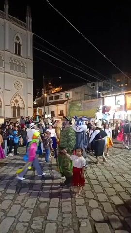 The video captures a nighttime street scene with a lively crowd of people, many dressed in colorful costumes and traditional attire. The scene takes place in a cobblestone-paved square, bordered by buildings of varying heights and styles. A tall, ornate church with arched windows stands prominently on the left side. The street is well-lit by overhead lights, casting shadows and illuminating the faces of the participants. People are dancing and moving around, contributing to a festive atmosphere. A flag with multiple colors is waving in the air, adding to the lively mood. Various characters, including one in military fatigues and a costumed mascot, interact within the crowd, engaging with children. The overall impression is that of a cultural celebration or festival.
