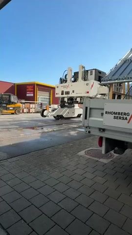 A person in an orange shirt walks across a paved area in front of a large piece of machinery attached to the back of a white truck. The machinery is white with some black and red accents and has a complex structure with multiple components. The truck has "RHOMBERG SERSA" written on its side. To the left, a yellow forklift is parked near a stack of brick-like objects. In the background, a yellow building with a red awning and a large garage door is visible. The sky is clear and blue. The ground is paved with grey interlocking bricks.