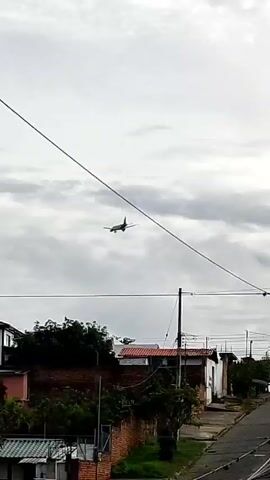 The video shows an overcast urban scene. Initially, a street lined with utility poles and wires is visible. Then, the camera pans upwards, showcasing clotheslines laden with laundry on rooftops. Finally, an airplane descends through the cloudy sky, appearing to approach a nearby airport situated amidst the buildings.