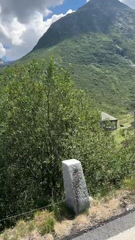 The video shows a mountainous landscape with a road in the foreground. The mountains are covered in lush green vegetation. There is a small building with a roof on the side of the mountain. The sky is cloudy. The camera appears to be moving, likely from a vehicle.