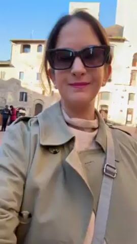 The video features a woman standing in a sunny outdoor location that appears to be a town square in Europe, possibly Italy. She is wearing a tan trench coat, a light-colored turtleneck sweater, and dark sunglasses. She has a crossbody bag slung over her shoulder. The background shows stone buildings with windows and doors, along with other people walking around in the distance. The camera is at eye level and remains mostly static, with slight movements likely due to the person holding it.