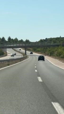 A vertical video shows a view from the passenger seat of a car driving on a highway. The car is traveling on a multi-lane highway with a clear blue sky and sparse vegetation visible on the sides. In the distance, there is a pedestrian overpass crossing the highway. Several cars are visible on the highway, some ahead and some in the opposite direction. The road appears to be in good condition, and the weather is sunny. The video captures the motion of the car as it navigates the highway, with the landscape blurring slightly due to the speed. The audio is a repetitive, electronic melody.
