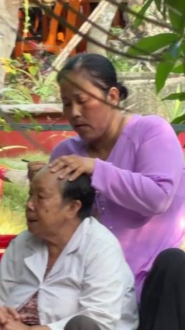 Two women are featured in this outdoor scene in Thoi Son, Vietnam. An older woman with very short hair or a partially bald scalp is seated, wearing a white shirt over a patterned top. She looks calm, gazing slightly to her left. Behind her, a younger woman in a long-sleeved purple top is standing and attending to the older woman's head. She holds her left hand on the older woman's head and uses a small, dark tool, possibly a comb or applicator, with her right hand, suggesting she is styling her hair or applying a treatment. The setting is a bright, sunny outdoor area with lush greenery, potted plants, and some orange chairs visible in the background, hinting at a garden or patio. Both women appear relaxed and focused on the activity.