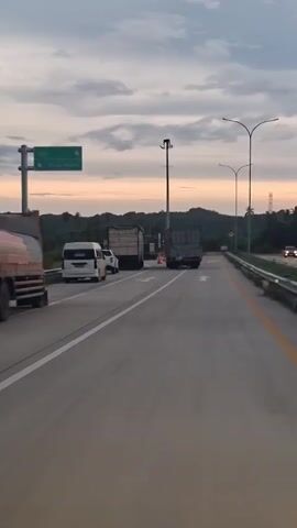 The video shows a highway with multiple lanes. Two large trucks are stopped on the right side of the road, with a white pickup truck and a white van further ahead on the left. A person in a blue shirt and black shorts is standing behind the second truck, seemingly interacting with its rear. The sky is overcast with hues of orange and purple, suggesting dawn or dusk. Streetlights and road signs are visible along the roadside. The overall atmosphere is calm, with no signs of distress or emergency.