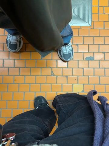 The video captures a first-person perspective looking down at two pairs of feet standing on a tiled floor with an orange grid pattern. The pair of feet in the foreground are wearing black shoes with white stripes and dark, loose-fitting jeans. A purple scarf or garment hangs from the side.  A pair of keys and a black object are visible near the bottom left. Another pair of feet, wearing light gray sneakers and blue jeans, are visible further away. A brown jacket or coat drapes down, obscuring the upper part of the person.  A metal grate is visible in the background.  A hand enters the frame holding a pair of glasses.