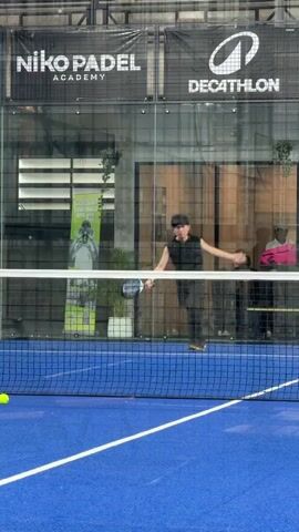 A young male in athletic wear plays padel. He is on a blue court, with a net separating him from the camera. The background shows a glass wall with advertisements for "Niko Padel Academy" and "Decathlon". The male hits a yellow ball with a black padel racket. The ball is in motion, as is the player. There are other yellow balls on the court. The lighting appears to be artificial, typical of an indoor sports facility.