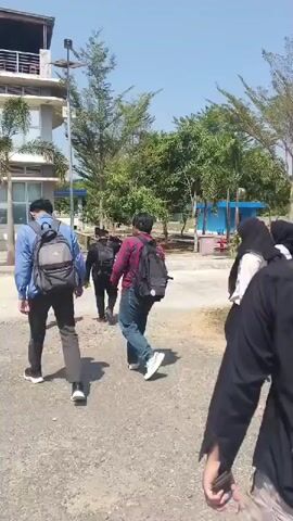 In the foreground, a person wearing a black hoodie and beige pants walks away from the camera. To their left, a person in a black hijab and black pants with a white shirt and black backpack walks towards the left side of the frame. To their left and slightly behind them, a person in a black hijab and black pants with a white shirt and a green object in their right hand walks away from the camera. Further left, a person in a dark colored shirt and dark pants with a black backpack walks away from the camera. Next to them, a person in a blue shirt and dark pants with a black backpack walks away from the camera. Behind them and to the left, a person in a white shirt and dark pants walks away from the camera. To the right of the person in the blue shirt, a person in a red and black plaid shirt and blue jeans with a black backpack walks away from the camera. In the background, there is a white building with balconies and windows, surrounded by trees and under a bright blue sky. The ground appears to be gravel and asphalt.