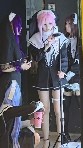 The media file depicts a group of individuals dressed in cosplay costumes resembling nuns, gathered in an indoor setting. The costumes are predominantly black and white, with some featuring colored wigs, such as purple and pink. One individual in the center, wearing a pink wig, is actively engaged with a smartphone mounted on a tripod, suggesting they are filming or streaming.

To the left, another individual with a purple wig is holding a smartphone and looking towards the central figure. In the foreground, a third person with a blue wig is kneeling and holding a pink drink cup. The background is dark, with some indistinct visual elements that could be posters or displays, hinting at a commercial or event space.

The lighting appears to be artificial, suggesting the scene is indoors and possibly at any time of day. The atmosphere seems casual and collaborative, with the individuals focused on their activity.

Visible text includes Chinese characters on what appears to be a poster in the background, though the text is not clearly legible. The pink cup also has some branding or logos, but these are not distinct enough to read. The overall scene suggests a cosplay gathering, possibly at an event or a dedicated photoshoot location in Shangcheng District, China. The attire and props indicate a focus on anime or manga-inspired characters.