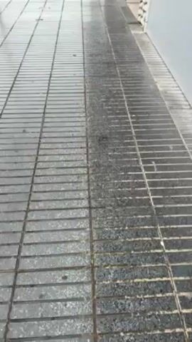 The video shows a wet, tiled sidewalk next to a street. Cars are driving on the wet road. In the background, there's a hill with colorful buildings. The sky is overcast. There are also streetlights and signs visible.