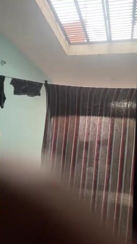 The video shows a dimly lit room with natural light filtering in from a skylight. Clothing is hanging on a clothesline. A striped curtain is visible. The camera appears to be moving, possibly handheld. A blurry foreground suggests the camera is being held by someone or is positioned close to an object.