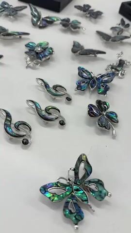 The video shows a selection of jewelry pieces laid out on a white surface. There are several butterfly-shaped brooches with iridescent blue and green coloring, along with dragonfly designs featuring similar coloring.  Additionally, there are a few brooches shaped like treble clefs, also featuring the iridescent material. The camera moves slightly, showing more of the jewelry spread across the surface.