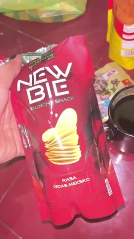 A hand is holding a red and black bag of "NEW BIE" crunchy snacks. The bag shows an image of potato chips and the text "RASA PEDAS MEKSIKO" which translates to "MEXICAN SPICY FLAVOR". The bag is partially opened, revealing snacks inside. To the right of the bag is a black mug filled with a dark liquid, presumably coffee or tea. Behind the mug is a yellow bag and a small packet with an image of sunflower seeds. Further to the right, a plate with a white and red rim holds four round biscuits and the mug of dark liquid. The surface appears to be a dark red, textured material.