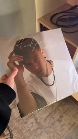 A person holds a record album in its original packaging. The album cover features a photo of a young man with short brown hair, wearing a white t-shirt and silver chain. Above the image is a handwritten signature, and the album title "Блокбастер". The back of the album lists the songs for Side A and Side B, and the year "2024 - 2025". The person turns the album to show both the front and back cover. The background shows a room with light grey walls, a wooden shelf, and black audio cables.