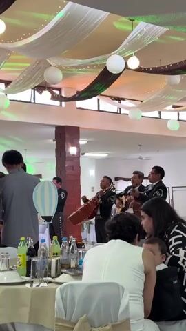 Four mariachi musicians dressed in black suits with embroidered accents are performing at what appears to be a celebration in a large indoor space. The musicians are playing their instruments and singing. One is playing a guitarron, two are playing guitars, and one is playing a wind instrument. They are surrounded by tables set for dining, with bottles of beverages and decorations. People are seated at the tables, listening to the music. The room is decorated with white fabric draped across the ceiling, interspersed with green accents, and round paper lanterns. A brick pillar stands near the musicians. The scene is well-lit with natural light coming through the windows and overhead lighting.