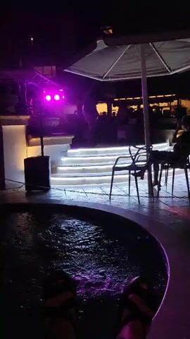 The video opens on a nighttime scene with a circular, dark-colored pool or jacuzzi in the foreground. A person's legs, wearing sandals, are visible near the edge of the pool, with ripples of water disturbed by their feet. Purple light emanates from the water.

In the mid-ground, there are several dark chairs and tables set up under a large, white umbrella. A person is seated at one of the tables. Beyond this area, there is a set of stairs illuminated with white lights, leading up to a raised platform.

On the platform, there are what appear to be stage lights, with two bright pink lights visible. Silhouettes of people are present on the platform, possibly a band or performers. In the background, there are blurred lights and structures, suggesting buildings or an establishment. The overall atmosphere is dimly lit and appears to be an outdoor entertainment venue at night.