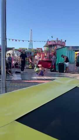 A group of people are at an outdoor amusement park. In the foreground, a trampoline with a yellow border is visible. In the background, several carnival rides and game booths are set up. A young man in a purple shirt and patterned shorts is breakdancing on a paved surface. He is performing a move where he is balancing on one hand with his legs extended. Another person in a black shirt stands nearby, observing. Several other individuals are scattered around the area, some watching the performance and others engaged in their own activities. The sky is clear and blue, suggesting a sunny day.