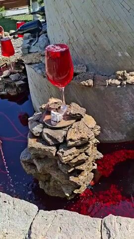 A close-up shot focuses on a fountain. In the foreground, a large wine glass filled with a red liquid is perched precariously on a stack of rocks. Beside it, another, smaller rock formation holds what appears to be a small silver box or container. The fountain itself is constructed with rough stones and a cylindrical structure in the background. The water in the basin is a deep red, with smaller red pebbles or stones scattered at the bottom. In the background, the fountain's design continues, with more large wine glasses similarly filled and placed on rock formations, suggesting a whimsical or artistic theme. The scene is outdoors, with sunlight casting shadows and highlighting the textures of the stones and the sheen of the liquid. The overall impression is one of an unusual, decorative fountain designed to resemble overflowing wine glasses.