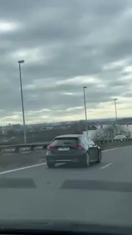 The video shows a view from inside a car driving on a highway on a cloudy day. Several other cars are visible on the road. On the right side of the highway, there is a river or body of water with bare trees on the banks. Streetlights line the side of the road. In the distance, a city skyline is visible. The camera remains steady throughout the video.