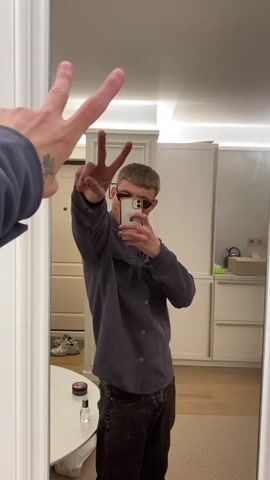 A person is taking a mirror selfie. They are wearing a dark grey shirt and dark pants and sunglasses. Their left hand is in the foreground, held up with two fingers extended. The person in the mirror is holding a phone and making a peace sign with their right hand. The background appears to be a room with white cabinetry and a door. There is a round table with some objects on it in the lower left corner.