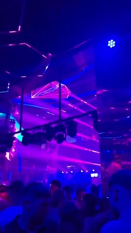 The video shows a crowded nightclub with blue and pink lights. People are dancing and socializing. Lights are projecting beams and patterns across the room. A large, mirrored structure is visible on the left side of the screen. Several spotlights are mounted on the ceiling.