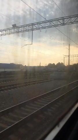 The video shows a view from a moving train, looking out the window at a train yard. The sky is cloudy with the sun setting, casting a warm glow. Multiple train tracks are visible, with some gravel ballast and ties. There are overhead power lines and poles for the electric trains. In the background, there are some industrial buildings and trees. The train appears to be moving at a moderate speed. The reflection of the train's interior is visible on the window.