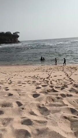 A video shot from a low angle on a sandy beach shows a body of water with a rocky outcrop in the distance. Several people are visible in the water, and footprints are scattered across the sand in the foreground. The sky is clear and overcast. The camera pans to the left and then to the right, revealing more of the coastline and the water.